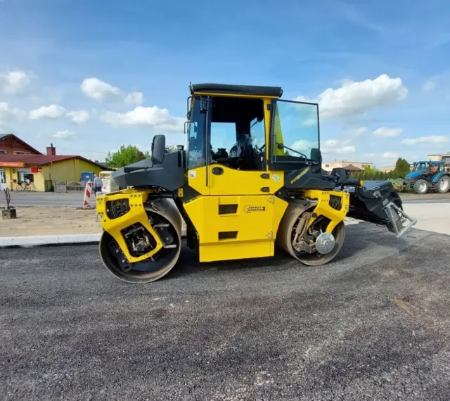 Żółty walec drogowy ugniata świeży asfalt. W tle widać budynki i niebieski traktor.