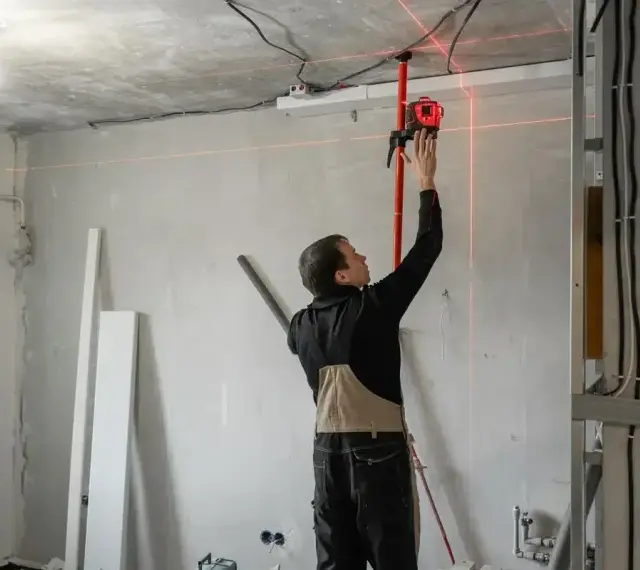 Budowlaniec ustawia laser, który wyznacza idealny pion murarski. Czerwone linie lasera przecinają pomieszczenie, pomagając w precyzyjnym montażu.