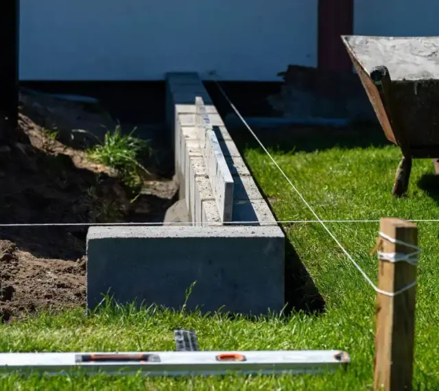 Budowa fundamentu pod ogrodzenie. Betonowe bloczki tworzą solidną podstawę, a sznur wyznacza linię. W tle taczka i zielona trawa.