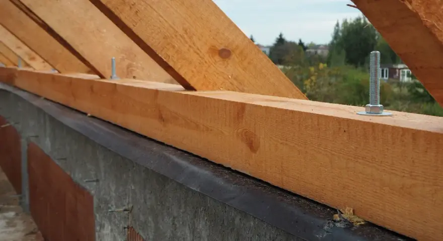 Drewniana murłata z widocznymi wymiarami, przykręcona do betonowej ściany. W tle drzewa i domy.