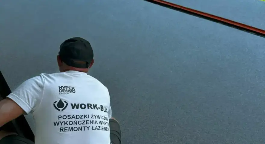 Pracownik w koszulce z napisem "Posadzki żywiczne" wykonuje prace na ciemnej, gładkiej powierzchni.