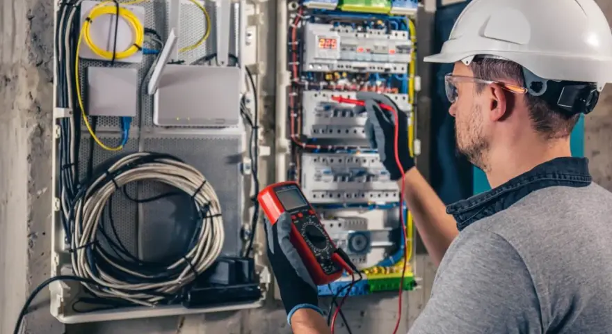 Elektryk w kasku przeprowadza przegląd instalacji elektrycznej, używając multimetru do pomiarów.