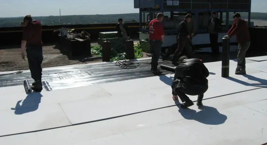 Mężczyźni pracują na dachu, układając materiał izolacyjny. W tle widać klimatyzator mgo.