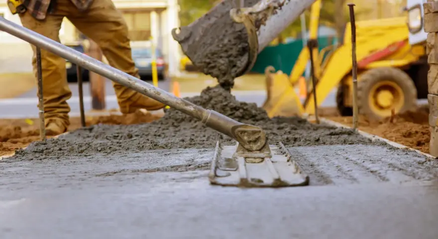 Pracownik rozprowadza świeży beton c12/15 za pomocą łaty. W tle widać koparkę i plac budowy.