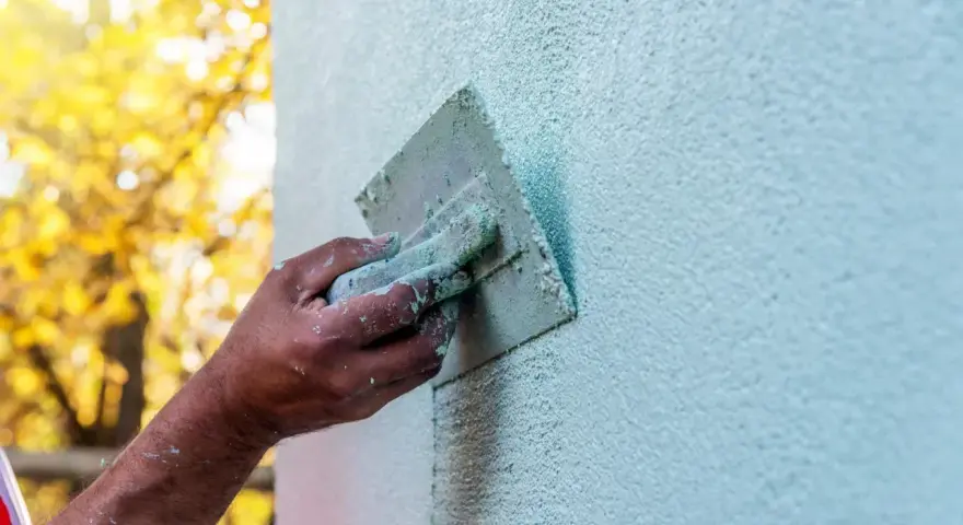 Ręka z kielnią wykonuje tynkowanie elewacji. Jasnozielony tynk pokrywa ścianę, tworząc fakturę. W tle jesienne drzewa.