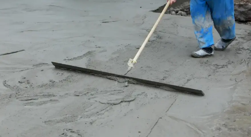 Pracownik w niebieskich ogrodniczkach wygładza świeży beton długą pacą. Trwa wygrzewanie posadzki.