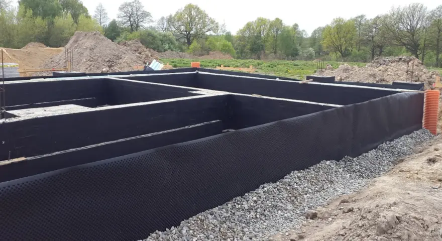 Fundament domu w trakcie budowy, zabezpieczony czarną membraną i otoczony żwirem.