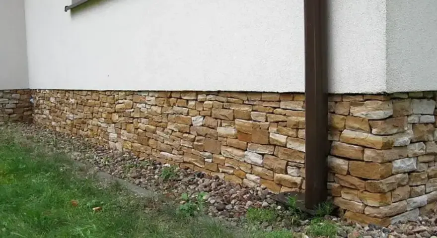 Eleganckie cokoliki z kamienia ozdabiają dom, tworząc naturalną, kamienną opaskę wokół białej ściany.