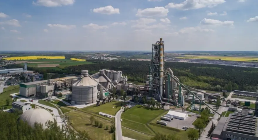Zakłady Lafarge z lotu ptaka, z dominującą wieżą pieca cementowego i silosami, otoczone zielenią i polami.