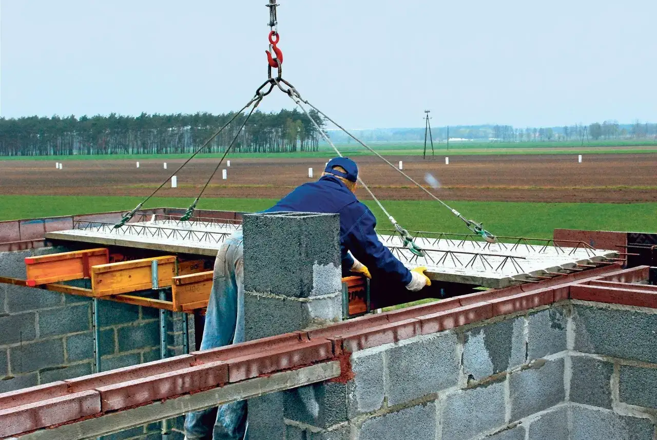 Robotnik z pomocą dźwigu montuje strop filigran na budowie. W tle pola i las.