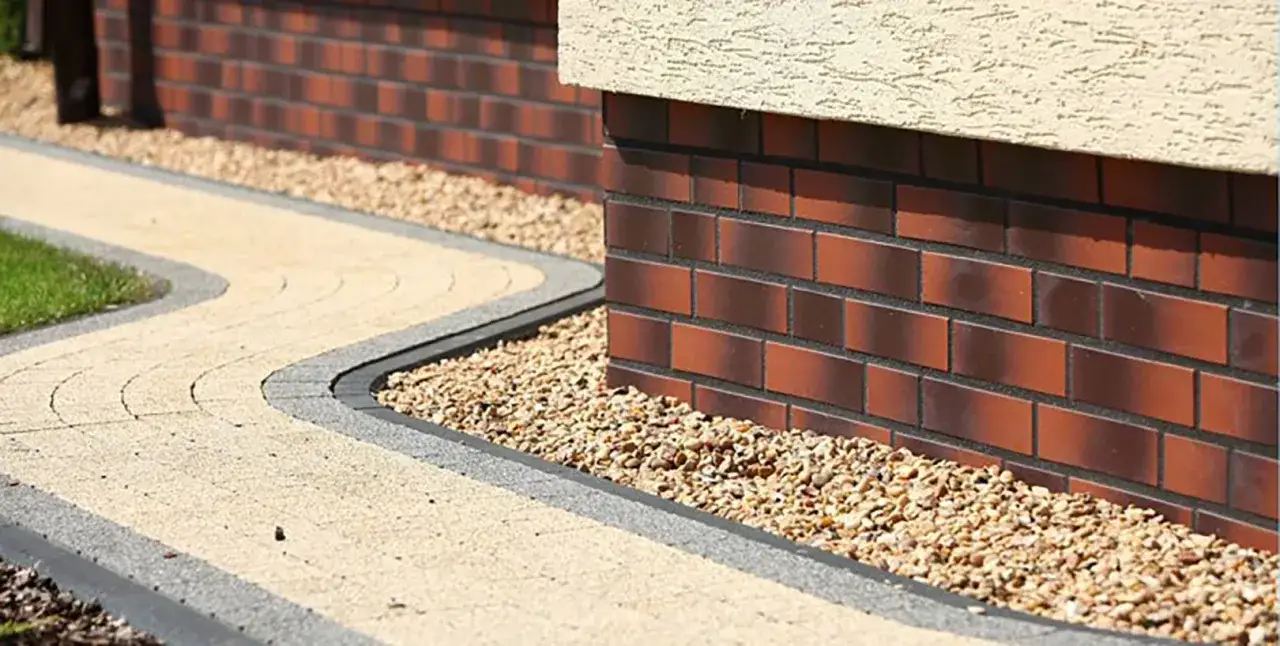 Elegancki cokół z płytek ceramicznych otacza budynek, obok kręta ścieżka z kostki brukowej i żwirowy pas.